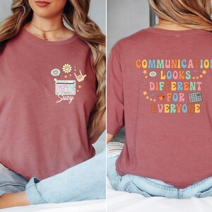 May include: A dusty rose t-shirt with the text "COMMUNICATION LOOKS DIFFERENT FOR EVERYONE" on the back. The front features a speech bubble, daisy, heart, hand sign, and a keyboard with the name "Suzy".