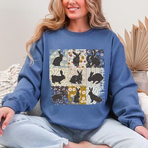 Puede incluir: Una sudadera azul con un dise&ntilde;o de patchwork. Presenta siluetas de conejos negros sobre cuadrados florales y estampados. La sudadera tiene cuello redondo y mangas largas.