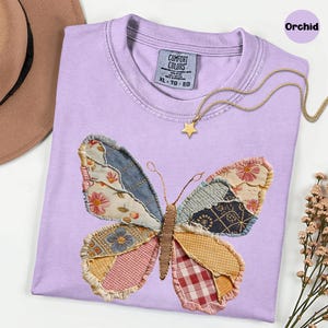 May include: A light purple Comfort Colors t-shirt with a patchwork butterfly design. The butterfly wings feature various fabric patterns and colors. A gold star pendant necklace rests on the shirt. The shirt size is XL-TG-EG.