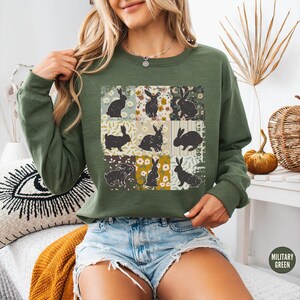 Puede incluir: Sudadera verde militar con un dise&ntilde;o de patchwork de siluetas de conejos negros sobre cuadrados florales y estampados. La sudadera tiene mangas largas y cuello redondo. Las palabras "MILITARY GREEN" est&aacute;n impresas en la parte inferior derecha.