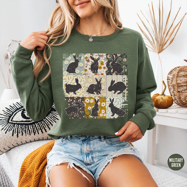 Puede incluir: Sudadera verde militar con un dise&ntilde;o de patchwork de siluetas de conejos negros sobre cuadrados florales y estampados. La sudadera tiene mangas largas y cuello redondo. Las palabras "MILITARY GREEN" est&aacute;n impresas en la parte inferior derecha.