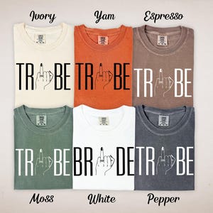 May include: Six different colored t-shirts, including ivory, yam, espresso, moss, white, and pepper. Each shirt has the word "TRIBE" printed in large, bold, white letters, with a hand graphic giving the middle finger.