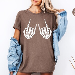 May include: A heather brown short-sleeve t-shirt featuring a white graphic print of two skeleton hands making a rock and roll hand gesture. The shirt has a crew neckline and a relaxed fit, with a small circular badge in the corner labeled Heather Brown.