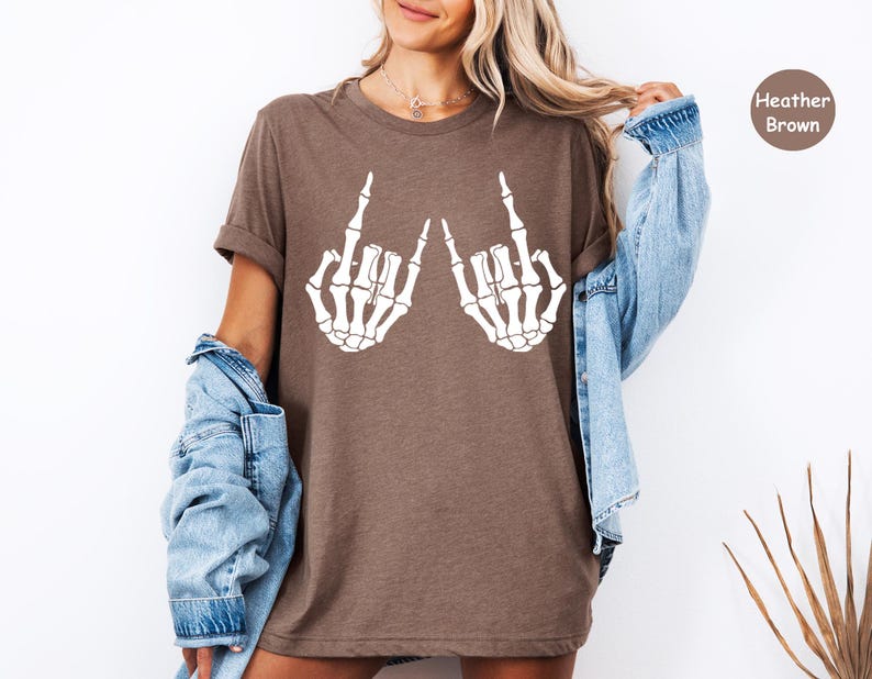 May include: A heather brown short-sleeve t-shirt featuring a white graphic print of two skeleton hands making a rock and roll hand gesture. The shirt has a crew neckline and a relaxed fit, with a small circular badge in the corner labeled Heather Brown.