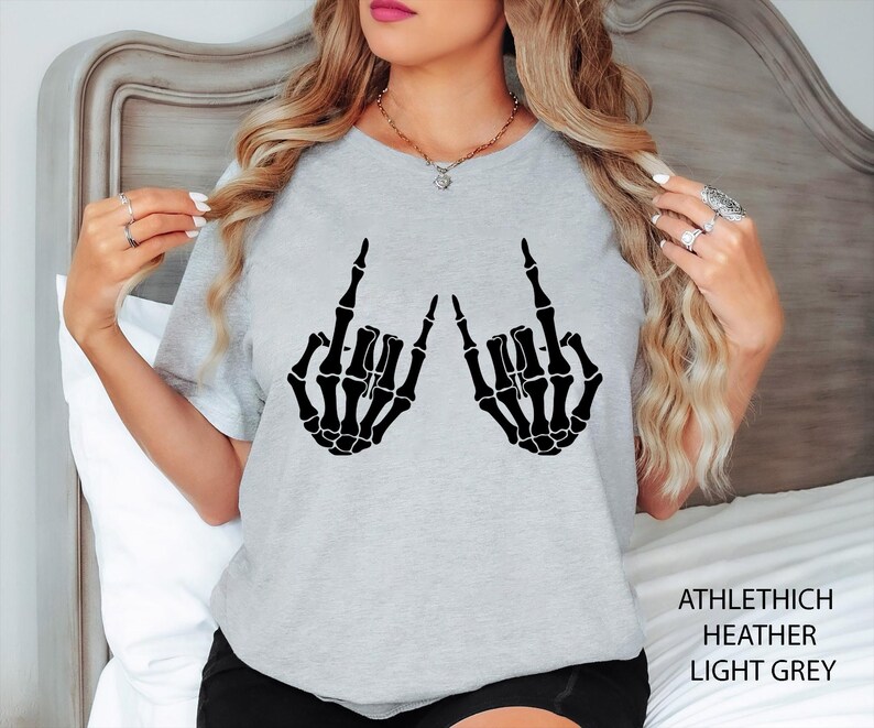 May include: A light grey t-shirt featuring a black graphic print of two skeleton hands making the rock and roll hand sign. The text ATHLETHICH HEATHER LIGHT GREY is printed in the bottom right corner of the image.