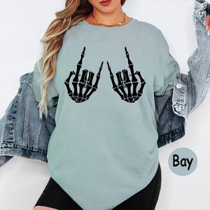 May include: A light blue crew neck t-shirt featuring a black graphic print of two skeleton hands making a rock and roll hand gesture. The shirt is layered under a blue denim jacket and paired with black shorts. The word Bay is printed in black.