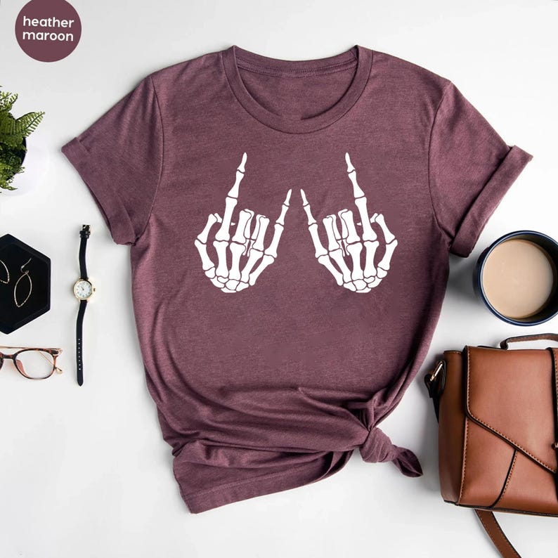 May include: A heather maroon short-sleeve t-shirt featuring a white graphic print of two skeleton hands making a rock and roll hand gesture. The shirt has a crew neckline and is displayed flat with the sleeves slightly rolled at the edges.