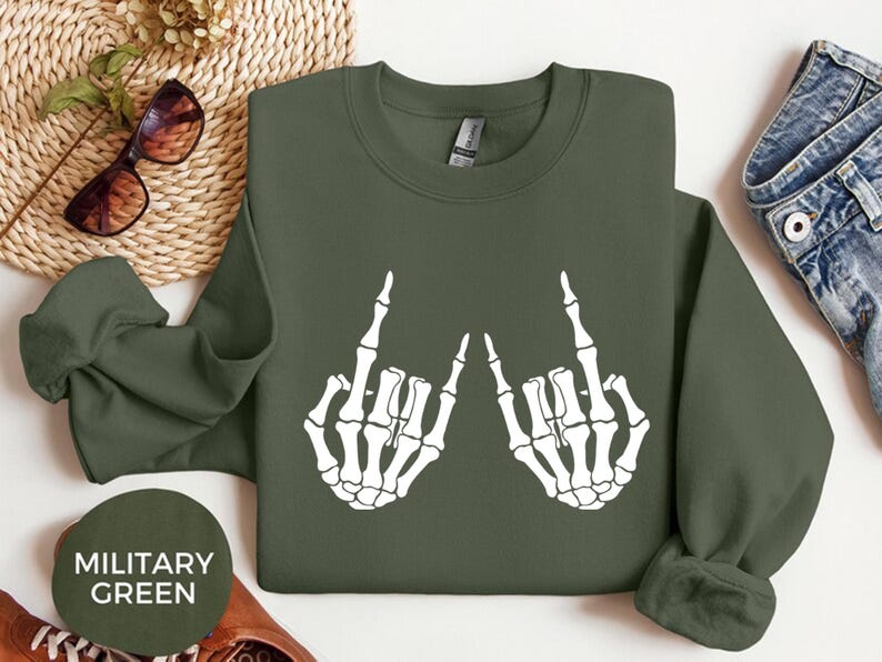 May include: A military green long-sleeve sweatshirt featuring a white graphic print of two skeleton hands making a rock and roll hand gesture. The sweatshirt has a crew neckline and long sleeves with ribbed cuffs, displayed flat with a circular label.