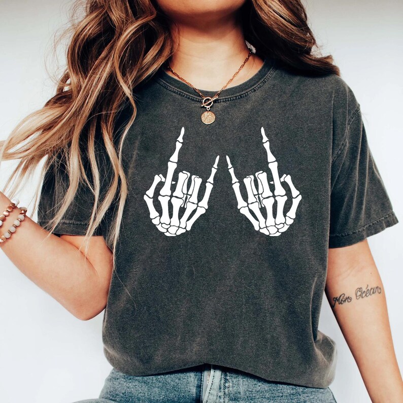 May include: A charcoal gray short-sleeve t-shirt featuring a white graphic print of two skeleton hands making the rock and roll hand gesture. The shirt has a crew neckline and a vintage-style washed fabric texture, providing a casual appearance.