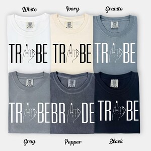 May include: Six different colored t-shirts are displayed, each with the word "TRIBE" and a hand with a ring on the middle finger. The colors are white, ivory, granite, gray, pepper, and black. The word "TRIBE" is in bold, capital letters.