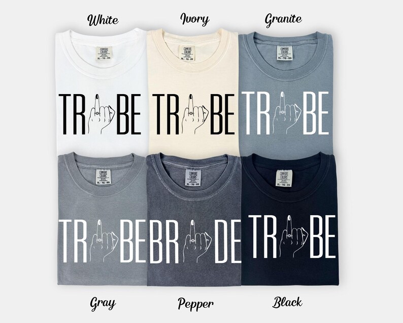 May include: Six different colored t-shirts are displayed, each with the word "TRIBE" and a hand with a ring on the middle finger. The colors are white, ivory, granite, gray, pepper, and black. The word "TRIBE" is in bold, capital letters.