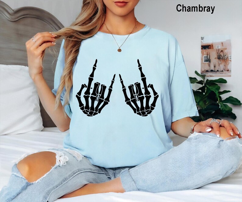 May include: A light blue t-shirt featuring a black graphic print of two skeleton hands making the rock and roll hand sign. The shirt has a crew neckline and short sleeves. The text Chambray is printed in the upper right corner of the image.