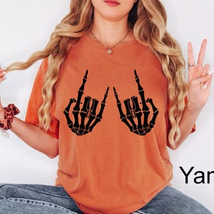 May include: A burnt orange short-sleeve t-shirt featuring a black graphic print of two skeleton hands making the rock and roll hand sign. The shirt has a crew neckline and a relaxed fit. The word Yam is printed in black in the bottom right corner.