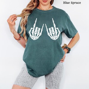 May include: A blue spruce coloured short-sleeve t-shirt featuring a white graphic print of two skeleton hands making a rock and roll hand gesture. The shirt has a crew neckline and a relaxed fit, with the text Blue Spruce printed in the corner.
