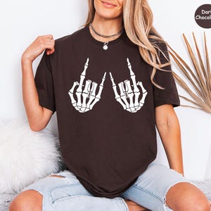 May include: A dark chocolate brown short-sleeve t-shirt featuring a white graphic print of two skeleton hands making the rock and roll hand sign. The upper right corner of the image contains a small circular badge with the text Dark Chocolate.