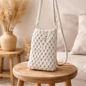 Könnte beinhalten: Eine cremefarbene Makramee-Umhängetasche mit gewebtem Muster, die an einem Riemen mit goldfarbenen Ringen hängt. Die Tasche wird auf einem kleinen Holzschemel gezeigt. Der Hintergrund umfasst eine Vase mit getrockneten Pflanzen und ein Sofa.
