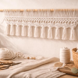 May include: A cream-coloured macrame wall hanging with a wooden dowel, featuring a geometric pattern and tassels. The image also shows yarn, a basket of yarn, and crafting supplies, suggesting a DIY home decor theme.