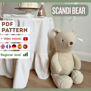 May include: A plush, beige teddy bear sits beside a table draped with a white tablecloth. The table holds a cake and a party hat. Text on the image reads "SCANDI BEAR" and "PDF PATTERN + Video tutorial".