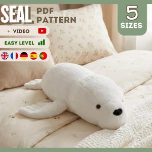 May include: A plush white seal toy with black eyes and a black nose is lying on a bed. The image includes text that says "SEAL PDF PATTERN", "+ VIDEO", "EASY LEVEL", and "5 SIZES".