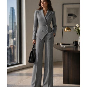 Op de afbeelding: Een grijze, dubbelbreasted pak met broek met wijde pijpen. Het pak heeft een getailleerde blazer met twee rijen knopen en een revers. Een zwarte handtas wordt in één hand gehouden. De achtergrond omvat een stadsgezicht zichtbaar door een groot raam.