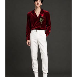 May include: A long-sleeved deep red velvet button-down shirt with a collar and a single chest pocket, paired with crisp white trousers, a matching white belt, and white leather ankle boots. A single red rose is tucked into the shirt pocket.