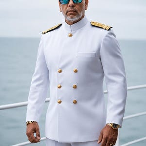 Pode incluir: Casaco de uniforme branco de abotoamento duplo com botões dourados e ombreiras. O casaco é combinado com calças brancas. A pessoa está usando óculos de sol azuis espelhados e um relógio e pulseira dourados. O fundo é uma massa de água.