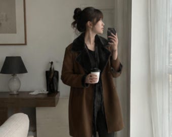 May include: A brown coat with a dark charcoal faux fur collar and interior lining. The coat features a double-breasted button closure, long sleeves, and a mid-thigh length. The outer material has a smooth, matte finish, while the dark fur trim is visible along the lapels and the inner edges of the garment.