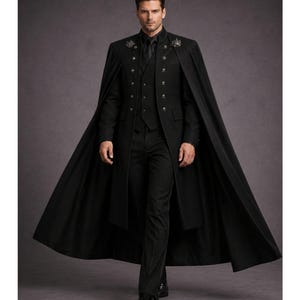 Op de afbeelding: Een zwarte formele jas met een cape, vest en broek. De jas heeft decoratieve knopen en versieringen op de schouders. De outfit is volledig zwart, wat een strakke en elegante uitstraling creëert. De cape van de jas voegt een dramatische flair toe.