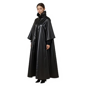 May include: Full-length black cape with a high collar. The cape has a flowing design and is made of a shiny, leather-like material. The model is wearing a black top and black shoes.