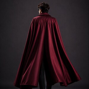 May include: A burgundy cape with a collar, flowing down to the ankles. The cape is made of a shiny, silky material. The person is wearing black pants and shoes. The background is a dark gray.