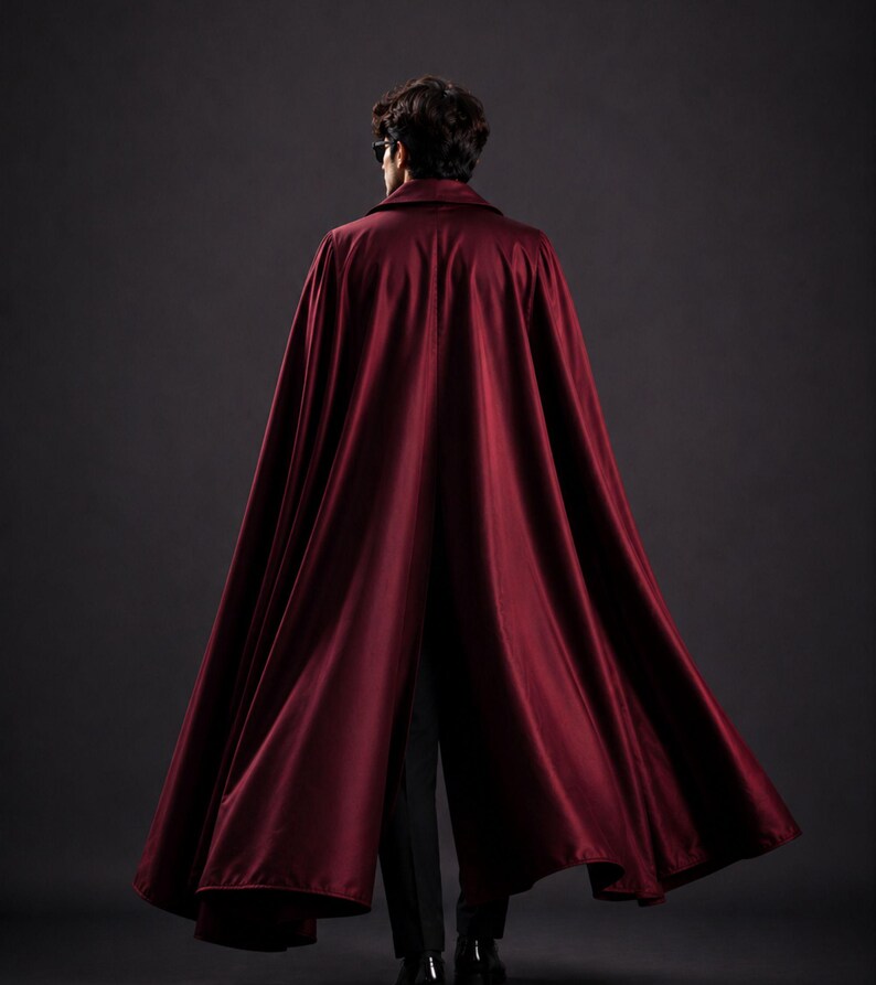 May include: A burgundy cape with a collar, flowing down to the ankles. The cape is made of a shiny, silky material. The person is wearing black pants and shoes. The background is a dark gray.