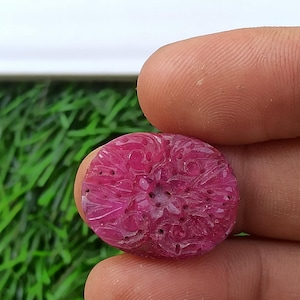 Könnte beinhalten: Ein geschnitzter, runder, rubinroter Edelstein. Der Edelstein hat ein detailliertes Blumenmuster, das in seine Oberfläche geschnitzt ist. Der Stein wird zwischen zwei Fingern vor einem Hintergrund aus grünem Gras und weißem Himmel gehalten.