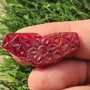 Könnte beinhalten: Ein herzförmiger, geschnitzter Edelstein in tiefroter Farbe. Die Oberfläche weist ein florales Design mit komplizierten Details auf. Der Edelstein wird zwischen den Fingern gehalten, mit grünem Laub im Hintergrund.