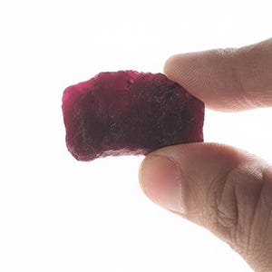 Könnte beinhalten: Ein grob geschliffener, dunkelroter Edelstein mit strukturierter Oberfläche. Der Rubin wird zwischen zwei Fingern vor weißem Hintergrund gehalten. Die Farbe des Edelsteins reicht von tiefem Burgunderrot bis zu einem helleren, leuchtenderen Rot.