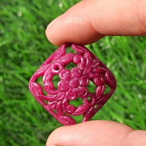 Könnte beinhalten: Ein geschnitzter, rubinroter Edelsteinanhänger mit floralem Design. Der quadratische Anhänger zeigt eine zentrale Blume, umgeben von filigranen Blatt- und Rankendetails. Der Edelstein wird zwischen zwei Fingern vor einem grünen Hintergrund gehalten.