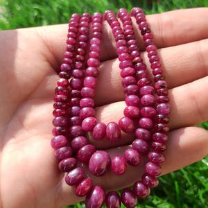 Könnte beinhalten: Eine Sammlung von Rubin-Halsketten. Die Halsketten bestehen aus kleinen, runden und ovalen Rubinperlen. Die Perlen haben eine tiefe, satte rote Farbe. Die Halsketten werden auf einer Hand vor einem Hintergrund aus grünem Gras präsentiert.