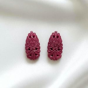 Könnte beinhalten: Ein Paar weinrote Ohrringe mit floralem Design. Jeder Ohrring hat eine geschnitzte, tropfenförmige Form mit aufwendigen Blumenmustern. Die Ohrringe werden auf einem weißen, seidigen Hintergrund präsentiert.