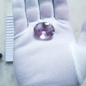 Könnte beinhalten: Ein oval geschliffener, facettierter Amethyst-Edelstein mit Lavendel- und Lilatönen liegt auf einem weißen Handschuh. Die Facetten des Edelsteins fangen das Licht ein und erzeugen einen funkelnden Effekt. Der Hintergrund ist neutral.