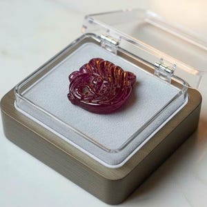 Op de afbeelding: Een gesneden, dieprode edelsteen in een doorzichtige vitrine. De edelsteen heeft een ingewikkeld ontwerp en is geplaatst op een grijs oppervlak in een houten omrande doos. De behuizing is open en onthult de details van de edelsteen.