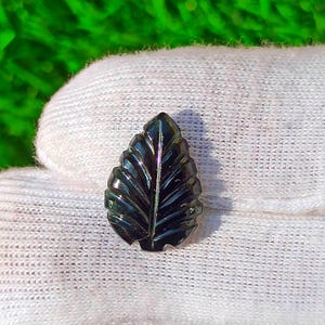 Könnte beinhalten: Ein dunkler, geschnitzter Edelstein in Form eines Blattes. Das Blatt hat detaillierte Adern und eine polierte, reflektierende Oberfläche. Der Edelstein ist ein tiefes, irisierendes Schwarz mit Andeutungen von Grün und Blau. Das Blatt liegt auf einer weißen Oberfläche mit grünem Hintergrund.