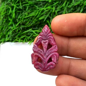 Könnte beinhalten: Ein geschnitzter Rubin-Edelsteinanhänger in Tropfenform. Der Anhänger hat ein aufwendiges Blumendesign mit filigranen Details. Der Rubin hat eine tiefrote Farbe und wird in einer Hand vor einem Hintergrund aus grünem Laub gehalten.