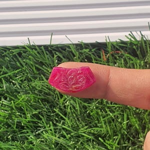 Könnte beinhalten: Ein facettierter, rosa Edelstein mit einem kunstvollen Blumendesign, das in seine Oberfläche geschnitzt ist. Der Stein wird zwischen Fingern gehalten, was seine polierten Facetten und seine lebendige Farbe zeigt. Der Hintergrund zeigt grünes Gras und eine weiße horizontale Oberfläche.