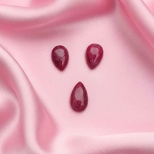 Könnte beinhalten: Drei tropfenförmige, tiefrote Edelsteine liegen auf einem rosa Satinstoff. Die Edelsteine haben eine polierte, reflektierende Oberfläche, und der Stoff hat weiche Falten. Die Edelsteine sind wahrscheinlich Rubine oder Granate.