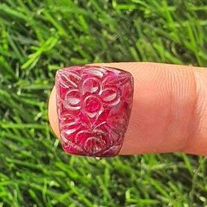 Könnte beinhalten: Ein rechteckiger, geschnitzter Rubin-Edelstein mit floralem Design. Der tiefrote Stein wird zwischen Fingern vor einem Hintergrund aus grünem Gras gehalten. Das geschnitzte Design zeigt eine zentrale Blume, umgeben von Blättern.