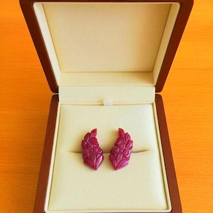 Könnte beinhalten: Ein Paar geschnitzte Rubinohrringe, präsentiert in einer offenen Holzschatulle. Die Ohrringe weisen ein florales Design auf und sind auf einem cremefarbenen Samtinnenraum platziert. Die Schatulle ist aus dunklem Holz mit goldfarbener Hardware gefertigt.