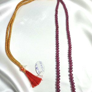 Könnte beinhalten: Eine Halskette mit einer Kette aus tiefroten, runden Perlen. Die Halskette hat eine gold-rote Kordel mit Quaste. Ein kleines Etikett mit Text ist an der Kordel befestigt.