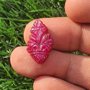 Könnte beinhalten: Ein geschnitzter, rubinroter Edelstein mit floralem Design. Der Edelstein weist eine detaillierte Schnitzerei einer stilisierten Blume und Blätter mit strukturiertem Sockel auf. Der Edelstein wird zwischen Fingern vor dem Hintergrund von grünem Gras gehalten.