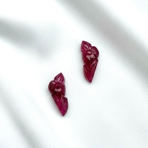 Puede incluir: Dos pendientes de flores de rubí tallado. Cada pendiente presenta un diseño floral detallado con pétalos y una flor central. Los pendientes son de color rojo intenso y están sobre un fondo blanco.