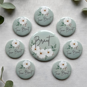 Könnte beinhalten: Eine Sammlung von runden, mintgrünen Buttons und einem größeren Button mit den Worten "Braut" und "Team Braut" in schwarzer Schrift. Jeder Button zeigt ein Aquarell-Blumenmuster mit weißen Blüten und grünen Blättern.