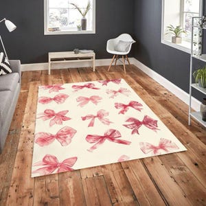 Può includere: Un tappeto rettangolare con sfondo color crema e un motivo di fiocchi rosa ad acquerello. I fiocchi, di varie dimensioni, sono sparsi sul tappeto. Il tappeto è posizionato su un pavimento in legno.